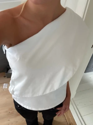 Vit oneshoulder topp med drapering - Säljer en vit oneshoulder topp med snygg draperad design. Toppen har en axel bar och är tillverkad i ett mjukt bomullsliknande material. Perfekt för dig som vill ha en unik och trendig look.