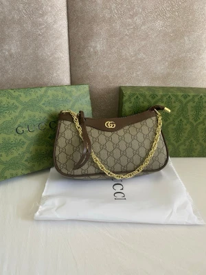 Gucci GG Supreme axelremsväska - Snygg axelremsväska från Gucci i beige GG Supreme canvas med bruna läderdetaljer. Väskan har guldfärgad kedja och GG-logo framtill. Klassisk halvmåneformad modell med dragkedja upptill och exklusiv känsla. Perfekt för dig som gillar lyxiga accessoarer.