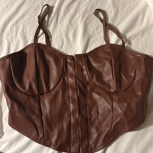 Brun läderkorsett med axelband - Snygg brun korsett i läderimitation med justerbara axelband och hakknäppning fram. Kuporna är formade för extra stöd och passform. Insidan har en mjukare beige yta för komfort. 
