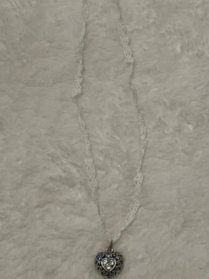 Halsband med hjärthänge och spetsband - Sött halsband med vitt spetsband och ett detaljerat hjärthänge i silverfärg. Hänget har ett mindre hjärta med glittrande sten i mitten och fina ornament runt om. Perfekt accessoar för dig som gillar romantisk och vintage-inspirerad stil.