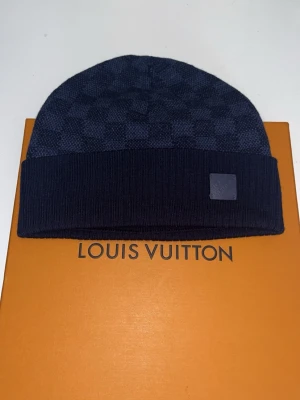 Mörkblå Louis Vuitton mössa ull - Snygg mörkblå mössa från Louis Vuitton i 100% ull med klassiskt rutmönster och ribbad kant. Märkets logotyp sitter diskret framtill på en liten patch. Perfekt accessoar för att hålla stilen under kalla dagar.