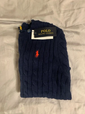Mörkblå kabelstickad tröja Polo Ralph Lauren - Mörkblå kabelstickad tröja från Polo Ralph Lauren med klassisk röd logga broderad på bröstet. Tröjan har rund halsringning, långa ärmar och ribbade muddar. Perfekt för dig som gillar stilrena och tidlösa plagg. Mått: axelbredd 35cm. Längd 60cm.