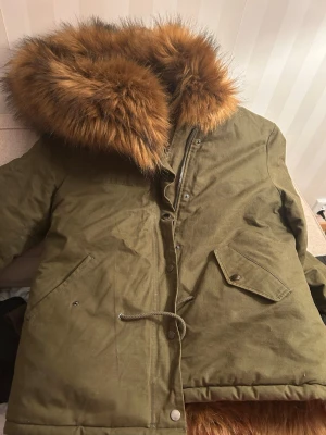 Olivgrön parkas med fuskpäls - Stilren olivgrön parkas med stor huva och fluffig brun fuskpäls runt kanten. Jackan har snörning i midjan, stora fickor med lock och tryckknappar. Perfekt för kalla dagar och ger en cool streetstyle-look. Använd ett par gånger bara! 