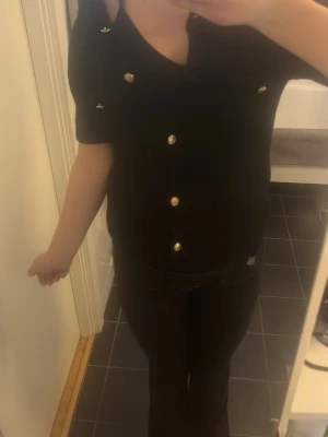 Svart topp med guldfärgade knappar - Snygg svart topp med korta ärmar och guldfärgade knappar framtill. Toppen har en v-ringad hals och dekorativa detaljer vid bröstet. Passformen är normal och materialet känns mjukt och stretchigt, perfekt till jeans eller kjol.