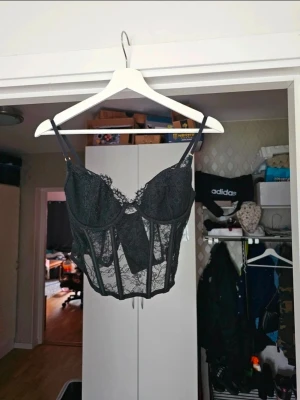 Svart spetskorsetttopp med gulddetaljer – H&M – Storlek 80B - Säljer en svart korsett topp med tunna axelband och vacker spetsdetalj. Modellen har snörning och hakar i ryggen, transparent mesh och blommönster i spetsen. Perfekt för att skapa en edgy look. Passar dig som gillar detaljer och trendiga toppar.