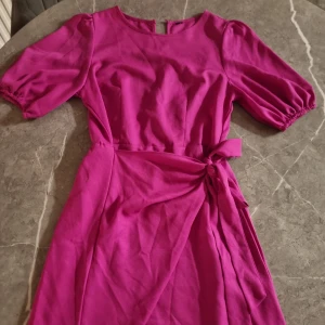 Rosa omlottklänning med knyt - Snygg rosa omlottklänning med knyt i midjan och puffiga korta ärmar. Klänningen har rund halsringning och en feminin siluett som framhävs av knytbandet. Perfekt för dig som vill ha en färgstark och trendig look.