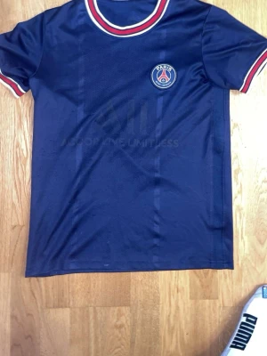 Blå PSG t-shirt med rödvita detaljer - Snygg blå t-shirt från Paris Saint-Germain med klubbens emblem på bröstet. Tröjan har rund hals och korta ärmar med rödvita ränder vid halsringning och ärmslut. Perfekt för dig som gillar fotboll och vill representera PSG.