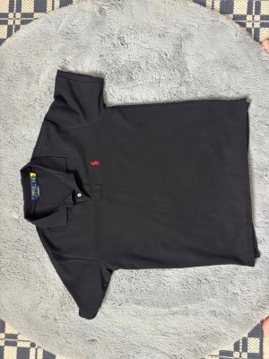 Svart pikétröja från Ralph Lauren - Klassisk svart pikétröja från Ralph Lauren med röd broderad logga på bröstet. Tröjan har korta ärmar, krage och två knappar vid halsen. Tillverkad i mjuk bomull som känns skön mot huden. Perfekt för en clean och stilren look.