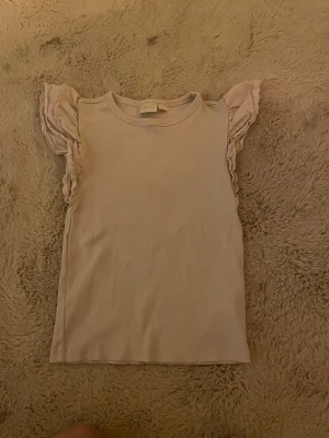 Rosa volangärmad topp Gina Tricot - Supersöt beige topp från Gina Tricot med volangdetaljer på ärmarna. Toppen är ribbad och har en enkel, rak passform. Perfekt för dig som gillar en feminin och stilren look.