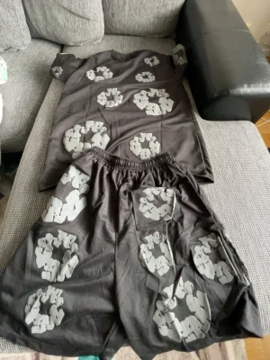 Svart set med grått mönster - Säljer ett svart set med t-shirt och shorts, båda plaggen har ett unikt grått mönster som liknar cirklar av figurer. T-shirten har rund hals och korta ärmar, shortsen har resår i midjan och avslappnad passform. Perfekt för dig som gillar statement-plagg.