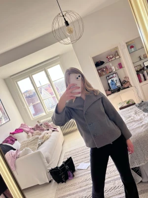 Grå kappa - Säljer en stilren grå kappa med stora knappar framtill och bred krage. Jackan har en croppad passform och är perfekt för dig som gillar en clean och modern look. Passar till både jeans och kjol för en trendig outfit.