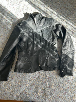 Svart skinnjacka med fickor - Snygg svart skinnjacka med dragkedja framtill, två bröstfickor med lock och två snedställda fickor med dragkedja. Jackan har markerade sömmar och förstärkningar vid armbågarna för en cool look. Figurnära passform och hög krage.