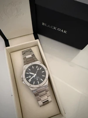 Silverfärgad klocka från Black Oak - Stilren klocka från Black Oak i silverfärgat metallarmband och boett. Urtavlan är svart med tydliga vita markeringar och visare. Klockan har en modern, kantig design och levereras i originalask. Nypris 799. Mitt pris 599. Använd 0 gånger eftersom den va för liten!!