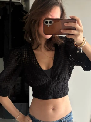 Svart croppad blus med puffärm - Trendig svart croppad blus med puffärmar och v-ringning. Toppen har ett rutigt transparent mönster och smockad midja som ger en snygg passform. Perfekt att styla med jeans eller kjol för en cool look.