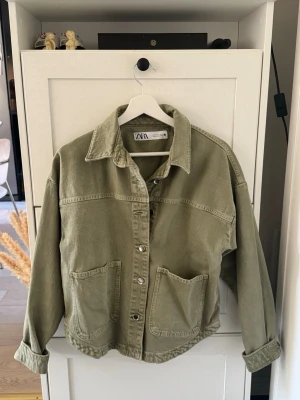 Olivgrön jeansjacka från Zara - Snygg olivgrön jeansjacka från Zara med avslappnad passform, stora framfickor och klassisk krage. Jackan har knäppning framtill och nedvikta ärmslut för en cool look. Perfekt att slänga över en hoodie eller t-shirt.
