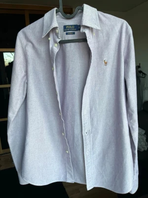  skjorta från Polo Ralph Lauren - Klassisk ljuslila skjorta från Polo Ralph Lauren i custom fit. Skjortan har lång ärm, knappar framtill och den ikoniska broderade loggan på bröstet. Tillverkad i mjuk bomull som känns skön mot huden. Perfekt för en clean och stilren look.
