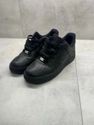  Nike Air Force 1  - Klassiska helsvarta Nike Air Force 1 sneakers i lågt utförande. Skorna har ovandel i slätt läder, perforerad tåbox för ventilation och kraftig sula. Swoosh-logga på sidan och metallbricka på snörningen. Perfekta för en clean och stilren look. Skorna är nästan helt nya och använda ett fåtal gånger, Skorna är köpta på zalando för 1300kr