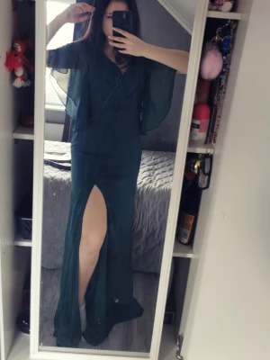 Mörkgrön långklänning med slits - Elegant mörkgrön långklänning med v-ringning, fladdriga korta ärmar och hög slits framtill. Klänningen har öppen rygg med knytband och är tillverkad i ett lätt, flytande material som ger en snygg siluett.