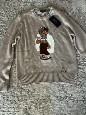 Beige stickad tröja bear - Stickad beige tröja med en bear. Perfekt för dig som gillar klassisk och lekfull stil. (Inte köpt från hemsidan). Helt ny. Har även i brun.