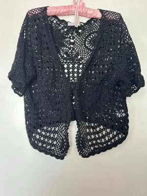 Svart virkad kofta från Fresh Point - Säljer en svart, kortärmad virkad kofta från Fresh Point. Koftan har ett öppet mönster med fina detaljer och en avslappnad passform. Perfekt att slänga över en topp för en cool look. Materialet är mjukt och luftigt.