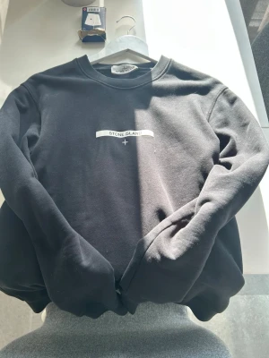 Svart sweatshirt från Stone Island - Snygg svart sweatshirt från Stone Island med rund halsringning och diskret logga framtill. Tröjan har en klassisk passform och är tillverkad i mjukt material som känns skönt mot huden. Perfekt för en clean och stilren look.