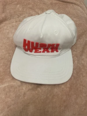 Vit keps från Huski Wear - Säljer en vit keps från Huski Wear med röd broderad text framtill. Klassisk sexpaneldesign med böjd skärm och ventilationshål. Tillverkad i bomullstyg för en bekväm och luftig känsla. Perfekt accessoar för en sportig och avslappnad stil.