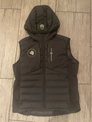 Svart dunväst från sail racing - Snygg svart dunväst från sail racing med huva och dragkedja framtill. Västen har ett coolt märke med Antarktis-tryck på bröstet och diskret logga längs dragkedjan. Perfekt för lager-på-lager och håller dig varm under kyliga dagar.