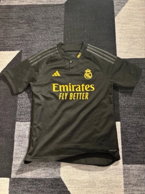 Custom Real Madrid Ronaldo matchtröja svart - Snygg svart Real Madrid matchtröja från Adidas med gula detaljer och tryck. Framsidan har klubbmärke och Emirates-logga, baksidan har Ronaldo och nummer 7 i gult. Tillverkad i lätt och ventilerande material, perfekt för fotbollsträning eller match. Custom 100% ekta