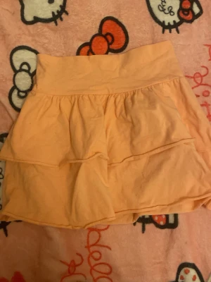Persikofärgad volangskort från H&M - Säljer en persikofärgad skort från H&M med dubbla volanger och bred midjeresår. Skorten har en mjuk och stretchig bomullskänsla och är perfekt för dig som gillar en söt och bekväm stil. Supersmidig att röra sig i och lätt att matcha med olika toppar.