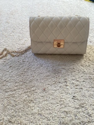 Vit quiltad axelväska med kedja - Stilren vit axelväska med quiltat mönster och guldfärgad kedjerem. Väskan har ett elegant lås i guld framtill och en klassisk rektangulär form. Perfekt för dig som gillar en tidlös och chic look.