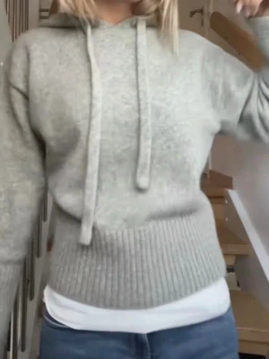 Ljusgrå hoodie med huva - Mysig ljusgrå hoodie med huva och dragsnören. Tröjan har ribbade muddar i nederkant och vid ärmslut, samt en avslappnad passform. Perfekt för en chill och stilren look. Materialet ser ut att vara mjukt och lite fluffigt, troligen en ull- eller bomullsblandning.