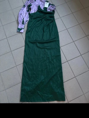 Grön långklänning med lila ärm by Malina  - Unik långklänning i grön satin med en axelbandslös design och en lång, puffig lila ärm med gröna detaljer. Klänningen har en asymmetrisk oneshoulder-look och är tillverkad i ett glansigt material som ger en lyxig känsla.