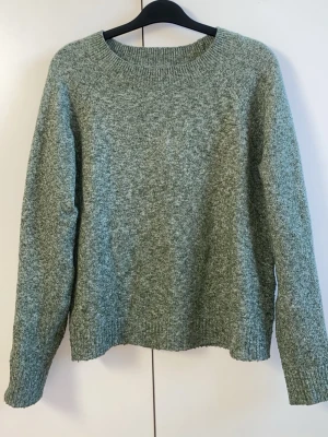 Grön stickad tröja från Vero Moda - Mysig grönmelerad stickad tröja från Vero Moda med rund halsringning och långa ärmar. Perfekt för lager på lager och enkel att matcha till jeans eller kjol. Ribbstickade muddar vid ärmslut och nederkant ger en snygg finish.