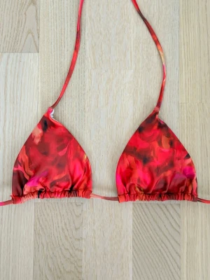 Röd och orange trekantsbikini - Snygg trekantsbikini i rött och orange med abstrakt mönster. Klassisk modell med knytband i nacken och ryggen. Materialet är troligtvis polyester eller nylon med stretch, perfekt för stranden eller poolen.