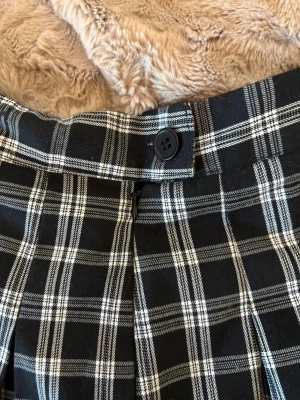Svartvit rutig kortkjol - Snygg svartvit rutig kortkjol med veckad design och knäppning framtill. Kjolen har klassiskt rutmönster och är perfekt för en trendig och ungdomlig stil. Passar till många olika outfits och är lätt att matcha med både sneakers och boots.