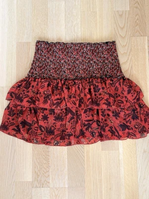 Röd volangkjol med blommönster - Kort kjol i rött med svart blommönster och smock i midjan. Kjolen har dubbla volanger och är gjord i ett lätt, luftigt tyg som ger en flowy känsla. Perfekt för dig som gillar boho-stil och vill ha något bekvämt och snyggt.