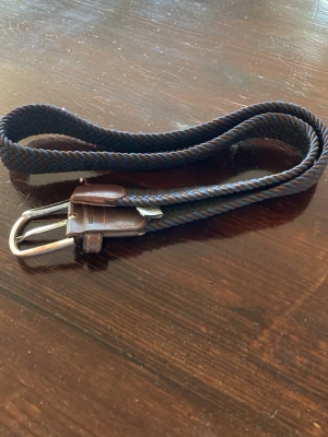 Mörkblått flätat bälte med skinn - Snyggt mörkblått flätat bälte från John Henric med detaljer i brunt skinn och silverfärgat spänne. Perfekt accessoar för att lyfta din outfit med en klassisk touch. Bältet har en flexibel design och passar till många olika stilar.