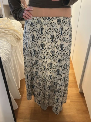 Vit långkjol med blått blommönster - Säljer en vit långkjol med blått blommigt mönster. Kjolen har en luftig och flowy passform, perfekt för varma dagar. Materialet känns lätt och bekvämt, och midjan är elastisk. Säljer för att den är för kort för mig!💗
