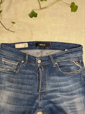 Blå Replay jeans med slitningar - Snygga blå jeans från Replay med klassisk femficksmodell och diskreta slitningar framtill. Jeansen har en rak passform och detaljer som logotyp på bakfickan och läderpatch i midjan. Perfekta för en avslappnad och trendig look.