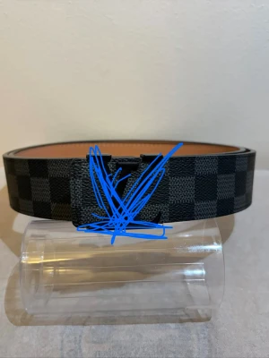 Svart och grått rutigt bälte - Snyggt bälte med svart och grått rutmönster och en matchande spänne framtill. Bältet har en stilren design och insidan är brun. Perfekt accessoar för att lyfta din outfit.