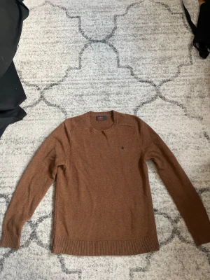 Brun stickad tröja från Morris - Snygg brun stickad tröja från Morris i 100% merinoull. Tröjan har rund halsringning, ribbade muddar och en liten broderad logga på bröstet. Perfekt för dig som gillar klassisk och stilren design.