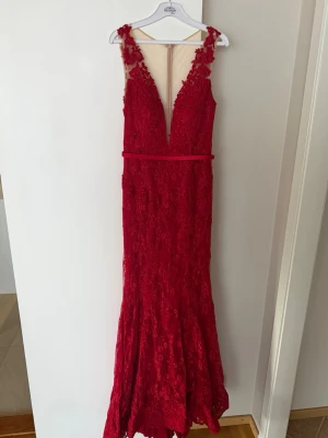 Röd långklänning med spetsdetaljer - Elegant röd långklänning från DRESSROOM med djup v-ringning och vackra spetsdetaljer. Klänningen har smala axelband, figurnära passform och dekorativa blommiga broderier med pärlor. Perfekt för bal eller festliga tillfällen. Aldrig använd. 