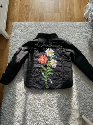 Svart jacka med blommor - Supercool handsydd quiltjacka med en blommig design. Jackan är svart, har olika blomstriga motiv, broderad med olika färg på garn i sömmarna. En one of a kind vårjacka!   Mått: Axel: 44cm Bröst: 54cm Längd: 63cm Armlängd: 60cm Jag på bilden bär vanligtvis storlek XS som referens.