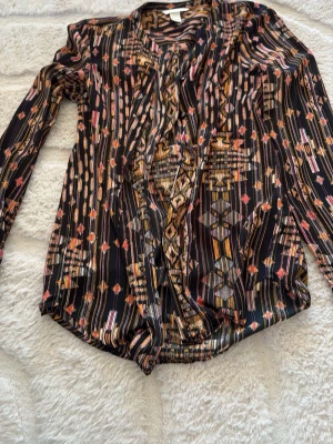 Mönstrad blus från H&M, svart/orange - Snygg blus från H&M i svart med färgstarkt etnoinspirerat mönster i orange, rosa och gult. Blusen har långa ärmar och ett lätt glansigt material som känns mjukt mot huden. Perfekt för dig som gillar unika prints och vill sticka ut.