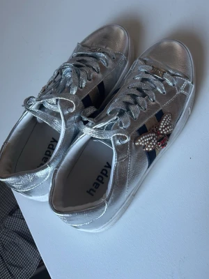 Silvriga sneakers med pärlbi - Snygga silvriga sneakers, blå och röda ränder på sidan samt en dekorerad pärlbi-detalj. Skorna har vita sulor och matchande silvriga skosnören. Perfekt för dig som vill sticka ut med coola detaljer och trendig look.