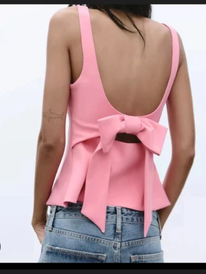 Rosa topp med rosett och öppen rygg - Supersöt rosa topp med fyrkantig urringning och breda axelband. Toppen har en stor rosett och öppen rygg som ger en unik look. Passar perfekt till jeans eller kjol för en trendig outfit.