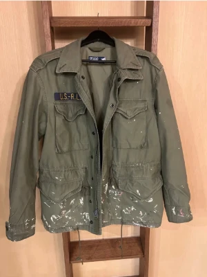 Ralph Lauren overshirt - Säljer en grön militärinspirerad overshirt från Polo Ralph Lauren med U.S.-RL-tryck på bröstet. Jackan har fyra fickor framtill, klassisk krage och stängs med dragkedja och knappar. Tillverkad i bomull och har vita färgstänk för en unik look.