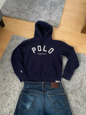 Mörkblå hoodie från Polo Ralph Lauren - Mörkblå hoodie från Polo Ralph Lauren med stor POLO-text och broderad logga på ärmen. Klassisk känguruficka framtill och ribbade muddar. Tillverkad i mjukt bomullsmaterial och har en skön huva. Perfekt för en avslappnad och sportig stil. (Passar M och S, jag har M och den passar mig rätt så bra👍🏼👍🏼)