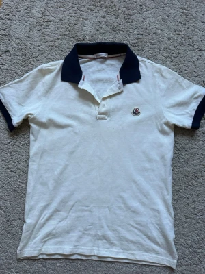 Vit Moncler pikétröja med mörkblå krage - Storlek 164cm vilket motsvarar xs kanske s. Klassisk vit pikétröja från Moncler med mörkblå krage och ärmslut. Tröjan har Moncler-logga broderad på bröstet och knäppning vid halsen. Perfekt för en stilren och avslappnad look.