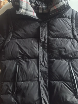 Svart jacka från Burberry - Snygg svart jacka från Burberry med patch på ärmen där det står 'BURBERRY LONDON ENGLAND'. Jacka har dragkedjor på fickorna och en avtagbar huva med rutigt foder i grått, vitt och rött. Perfekt för lager på lager och streetstyle.